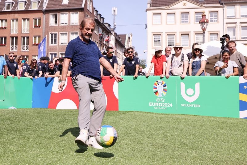 [{"clang_id":1,"value":"Mann kickt Fußball auf grünem Rasen. Im Hintergrund eine grüne Werbeplane mit UEFA Euro 2024 Logo und Menschen. Gebäude im Hintergrund."}]