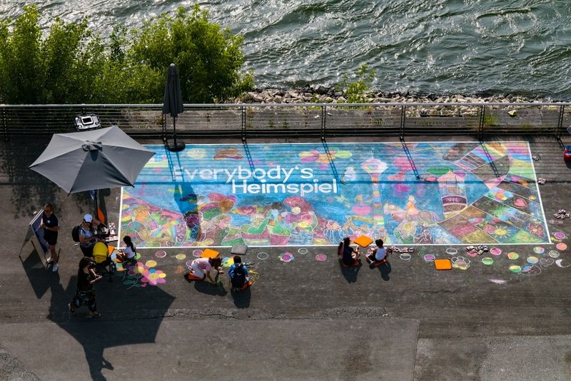 [{"clang_id":1,"value":"Luftaufnahme zeigt bunte Straßenkreidekunst auf einem Platz am Flussufer. Menschen sitzen oder knien um die Zeichnung herum, ein Sonnenschirm spendet Schatten."}]