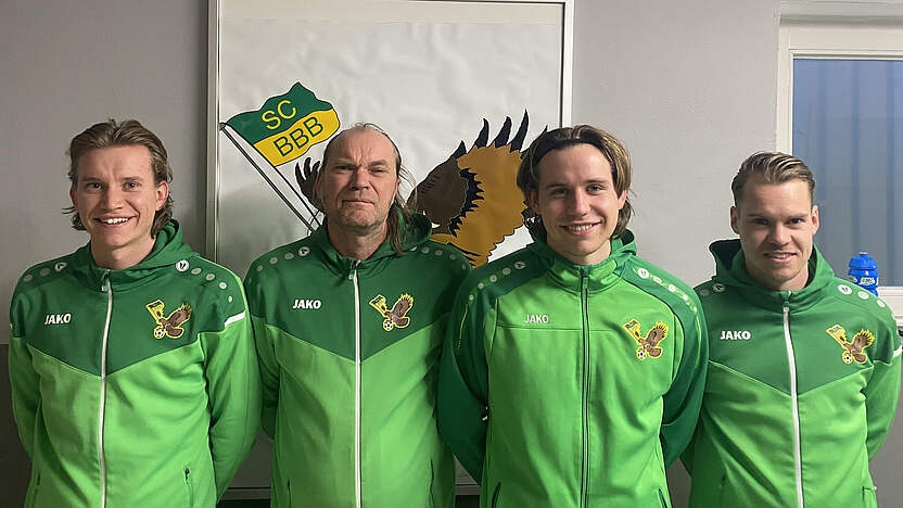 [{"clang_id":1,"value":"Vier Männer in grünen Trainingsanzügen mit Logo \"SC BBB\" stehen nebeneinander vor einer weißen Wand mit Vereinswappen."}]