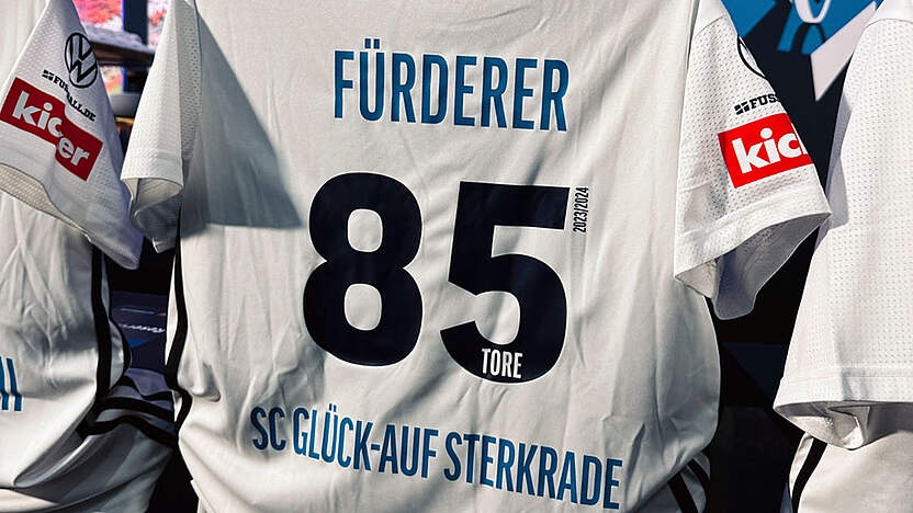 [{"clang_id":1,"value":"Fußballtrikot mit Namenszug \"FÜRDERER\", Nummer 85 und Schriftzug \"SC GLÜCK-AUF STERKRADE\". Trikot hängt an Bügeln, Sponsorenlogos sichtbar."}]