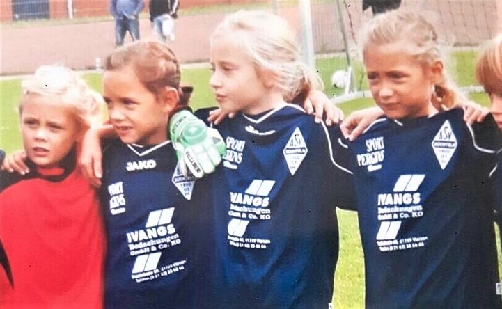 [{"clang_id":1,"value":"Fünf blonde Kinder stehen eng zusammen auf einem Fußballfeld. Vier tragen blaue Fußballtrikots mit Werbeaufdruck, eines ein rotes T-Shirt."}]