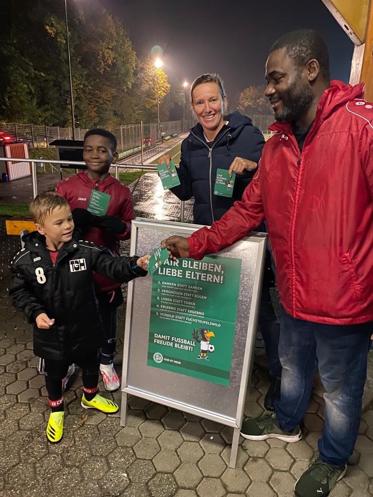 [{"clang_id":1,"value":"Drei Personen neben Info-Stand. Links Kind im Fußballtrikot, Mitte Frau mit Jacke, rechts Mann mit Jacke. Im Hintergrund Fußballfeld und Beleuchtung."}]