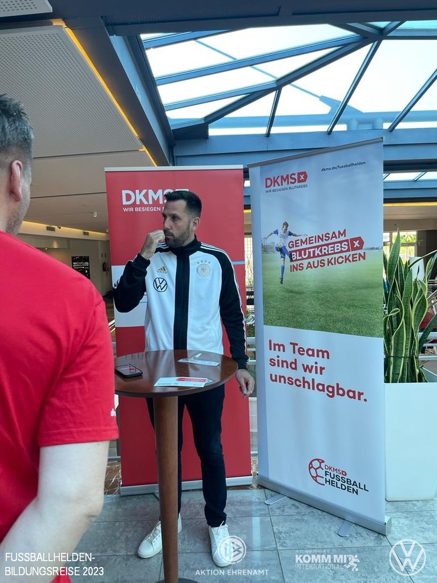[{"clang_id":1,"value":"Mann in Fußballtrikot vor großem Banner mit Aufschrift \"Gemeinsam Blutkrebs auskicken\". Im Hintergrund moderne Hotelhalle mit Pflanzen. Logos von DKMS und Volkswagen."}]