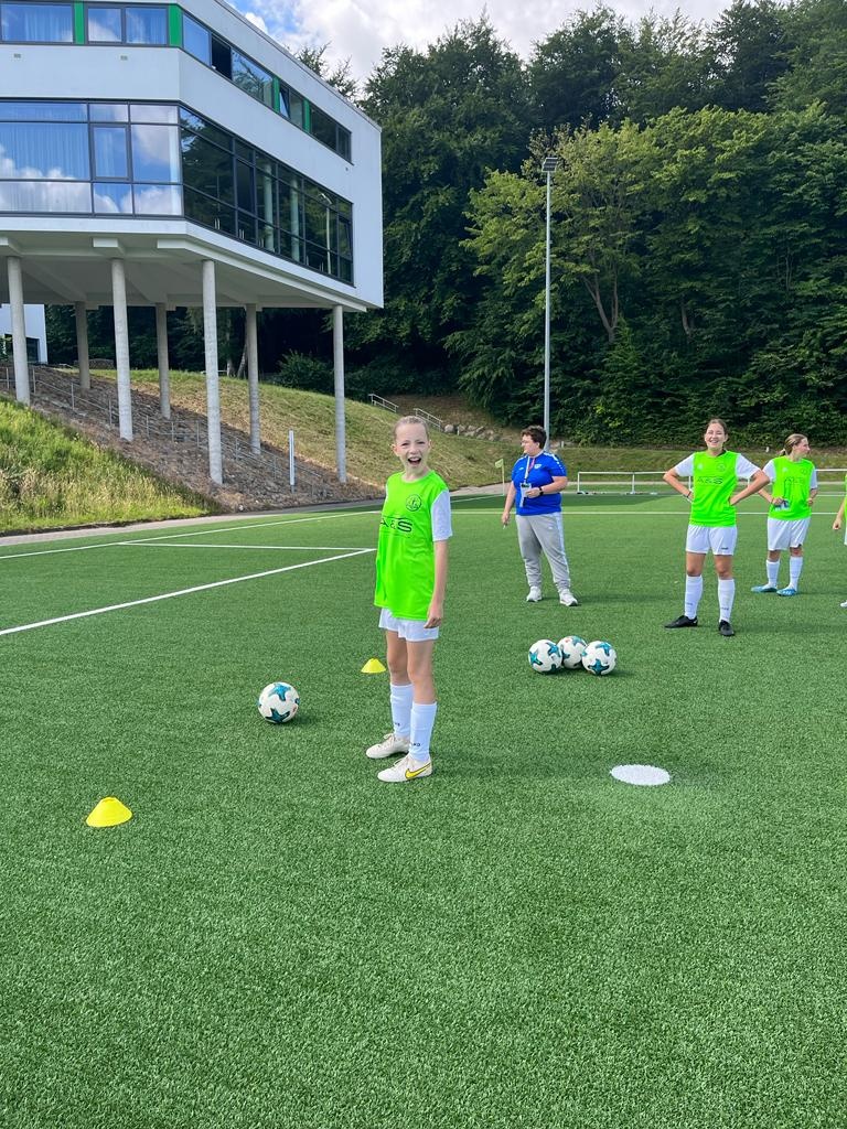 [{"clang_id":1,"value":"Fußballfeld mit Jugendlichen in Sportkleidung. Im Hintergrund ein modernes Gebäude mit Glasfassade und Grünanlagen. Mehrere Fußbälle und Hütchen auf dem Feld."}]