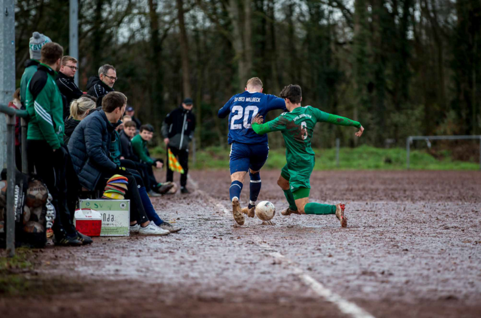 [{"clang_id":1,"value":"Fußballspiel im Freien. Zwei Spieler in Shorts und Trikots kämpfen um den Ball auf einem schlammigen Feld, Zuschauer am Spielfeldrand."}]