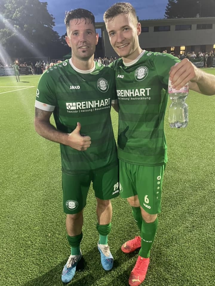 [{"clang_id":1,"value":"Zwei Fußballspieler in grünen Trikots und Shorts posieren lächelnd auf einem Fußballfeld. Spieler links zeigt Daumen hoch, Spieler rechts den Peace-Zeichen."}]