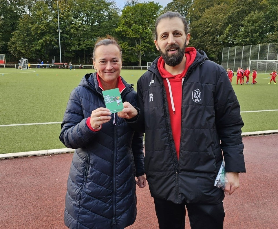 [{"clang_id":1,"value":"Zwei Personen, eine Frau mit blauem Jackett und ein Mann mit rotem Shirt und Trainingsjacke, stehen lächelnd vor einem Fußballfeld."}]