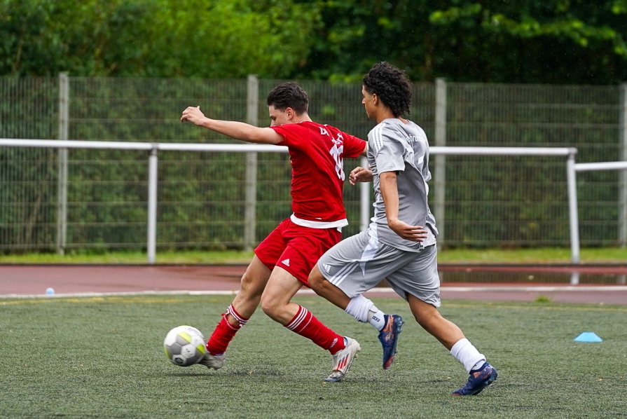 [{"clang_id":1,"value":"Zwei Fußballspieler im Wettkampf. Ein Spieler in Rot schießt auf das Tor, während ein Spieler in Grau ihn verfolgt. Hinter ihnen ein Zaun und Bäume."}]