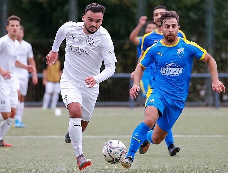 [{"clang_id":1,"value":"Zwei Fußballspieler im Wettkampf. Links Spieler mit weißem Trikot, Nummer 19, dribbelt den Ball. Rechts Gegenspieler in blauem Trikot, Nummer 15. Hintergrund weitere Spieler und Bäume."}]
