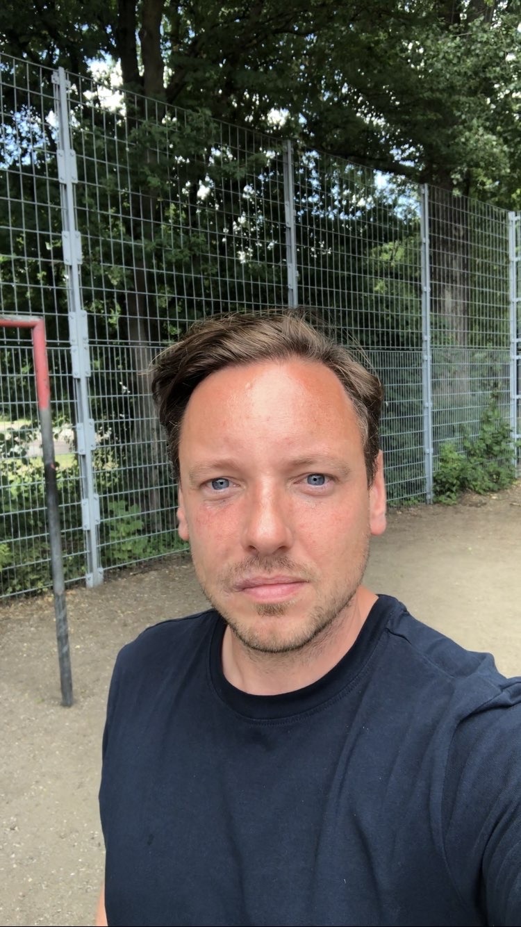 [{"clang_id":1,"value":"Mann mit braunen Haaren und blauem Blick vor Metallzaun und Bäumen. Er trägt ein dunkles T-Shirt."}]