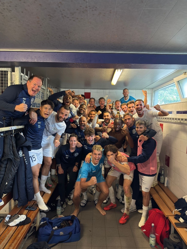 [{"clang_id":1,"value":"Fußballmannschaft in Umkleidekabine feiert. Spieler sitzen und stehen dicht aneinander, lächeln und jubeln. Dunkelblaue und weiße Sportkleidung. Holzbank und Schränke im Hintergrund."}]